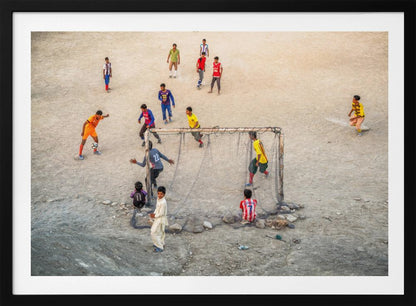 An overhead view of a group of boys and young men playing a spirited game of soccer on a large, dusty dirt field. A player in a bright orange uniform prepares to kick the ball towards a makeshift goal, defended by a goalie in a dark blue jersey. Other players in various colored shirts are scattered across the field, while a few young spectators watch from the sidelines. The entire image is enclosed in a silver frame. Wall Art