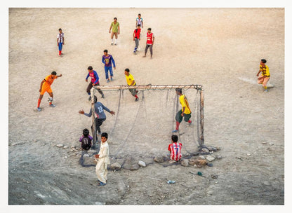 An overhead view of a group of boys and young men playing a spirited game of soccer on a large, dusty dirt field. A player in a bright orange uniform prepares to kick the ball towards a makeshift goal, defended by a goalie in a dark blue jersey. Other players in various colored shirts are scattered across the field, while a few young spectators watch from the sidelines. The entire image is enclosed in a silver frame. Wall Art