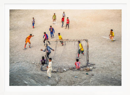 An overhead view of a group of boys and young men playing a spirited game of soccer on a large, dusty dirt field. A player in a bright orange uniform prepares to kick the ball towards a makeshift goal, defended by a goalie in a dark blue jersey. Other players in various colored shirts are scattered across the field, while a few young spectators watch from the sidelines. The entire image is enclosed in a silver frame. Wall Art