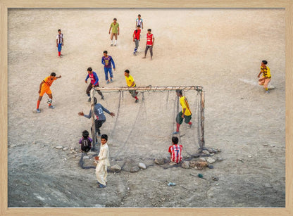 An overhead view of a group of boys and young men playing a spirited game of soccer on a large, dusty dirt field. A player in a bright orange uniform prepares to kick the ball towards a makeshift goal, defended by a goalie in a dark blue jersey. Other players in various colored shirts are scattered across the field, while a few young spectators watch from the sidelines. The entire image is enclosed in a silver frame. Wall Art