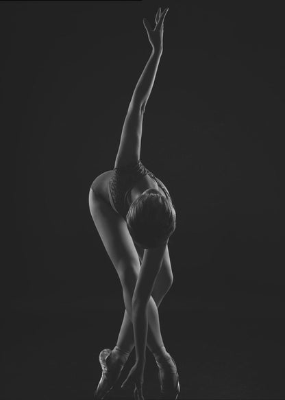 A dramatic black and white photograph of a ballerina posed against a dark background. She is bent forward at the waist, with her legs crossed and on pointe. One arm reaches up towards the ceiling in a graceful curve while the other hand touches the floor near her feet, showcasing her flexibility and strength. Wall Art