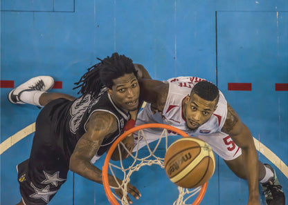 An overhead, bird's-eye view photograph of two basketball players looking up from below the hoop as a basketball goes through the net. The court is bright blue, and the image is enclosed in a silver frame. Poster
