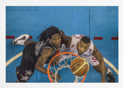 An overhead, bird's-eye view photograph of two basketball players looking up from below the hoop as a basketball goes through the net. The court is bright blue, and the image is enclosed in a silver frame. Poster