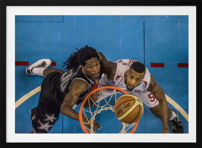An overhead, bird's-eye view photograph of two basketball players looking up from below the hoop as a basketball goes through the net. The court is bright blue, and the image is enclosed in a silver frame. Poster