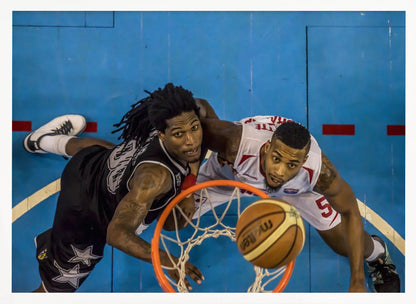 An overhead, bird's-eye view photograph of two basketball players looking up from below the hoop as a basketball goes through the net. The court is bright blue, and the image is enclosed in a silver frame. Poster
