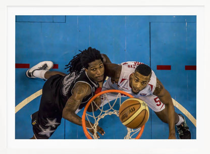 An overhead, bird's-eye view photograph of two basketball players looking up from below the hoop as a basketball goes through the net. The court is bright blue, and the image is enclosed in a silver frame. Poster