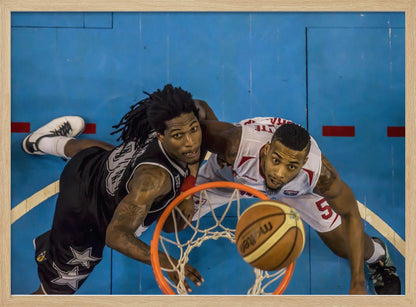 An overhead, bird's-eye view photograph of two basketball players looking up from below the hoop as a basketball goes through the net. The court is bright blue, and the image is enclosed in a silver frame. Poster