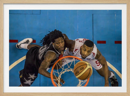 An overhead, bird's-eye view photograph of two basketball players looking up from below the hoop as a basketball goes through the net. The court is bright blue, and the image is enclosed in a silver frame. Poster