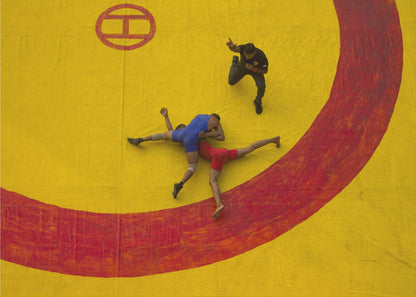 An aerial, high-angle photograph of a wrestling match in a silver frame. Two wrestlers, one in a blue singlet and the other in red, are locked in a hold on a vibrant yellow mat marked with a thick, painted red circle. A referee dressed in black stands nearby, gesturing. Artwork