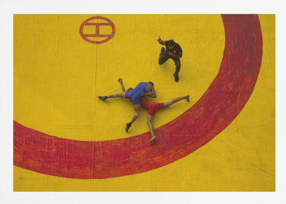 An aerial, high-angle photograph of a wrestling match in a silver frame. Two wrestlers, one in a blue singlet and the other in red, are locked in a hold on a vibrant yellow mat marked with a thick, painted red circle. A referee dressed in black stands nearby, gesturing. Artwork