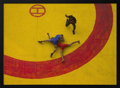 An aerial, high-angle photograph of a wrestling match in a silver frame. Two wrestlers, one in a blue singlet and the other in red, are locked in a hold on a vibrant yellow mat marked with a thick, painted red circle. A referee dressed in black stands nearby, gesturing. Artwork