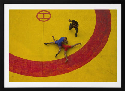 An aerial, high-angle photograph of a wrestling match in a silver frame. Two wrestlers, one in a blue singlet and the other in red, are locked in a hold on a vibrant yellow mat marked with a thick, painted red circle. A referee dressed in black stands nearby, gesturing. Artwork