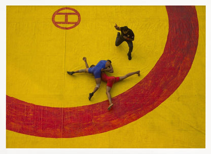 An aerial, high-angle photograph of a wrestling match in a silver frame. Two wrestlers, one in a blue singlet and the other in red, are locked in a hold on a vibrant yellow mat marked with a thick, painted red circle. A referee dressed in black stands nearby, gesturing. Artwork
