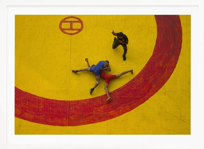An aerial, high-angle photograph of a wrestling match in a silver frame. Two wrestlers, one in a blue singlet and the other in red, are locked in a hold on a vibrant yellow mat marked with a thick, painted red circle. A referee dressed in black stands nearby, gesturing. Artwork