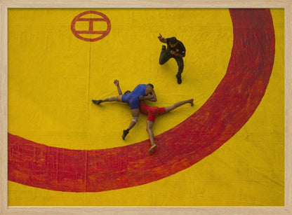 An aerial, high-angle photograph of a wrestling match in a silver frame. Two wrestlers, one in a blue singlet and the other in red, are locked in a hold on a vibrant yellow mat marked with a thick, painted red circle. A referee dressed in black stands nearby, gesturing. Artwork