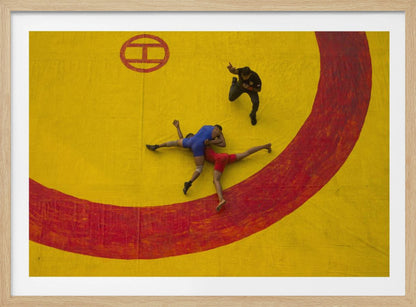 An aerial, high-angle photograph of a wrestling match in a silver frame. Two wrestlers, one in a blue singlet and the other in red, are locked in a hold on a vibrant yellow mat marked with a thick, painted red circle. A referee dressed in black stands nearby, gesturing. Artwork