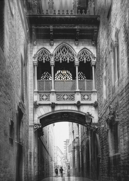 A moody, vertical black and white photograph of an ornate Gothic-style bridge connecting two stone buildings over a narrow alleyway. Below the arch of the bridge, the wet street stretches into the distance where two figures are walking away. Decor