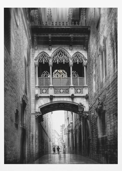 A moody, vertical black and white photograph of an ornate Gothic-style bridge connecting two stone buildings over a narrow alleyway. Below the arch of the bridge, the wet street stretches into the distance where two figures are walking away. Decor