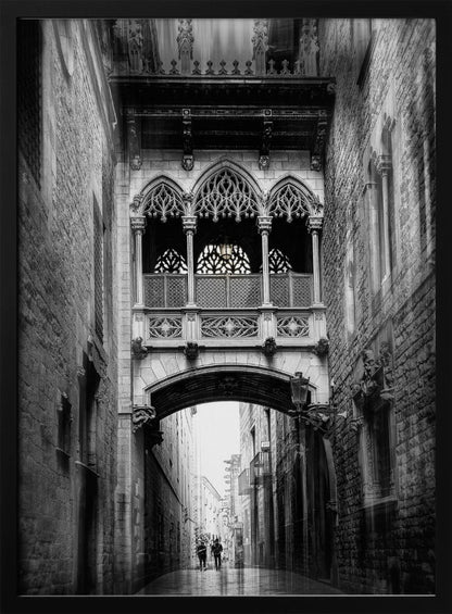 A moody, vertical black and white photograph of an ornate Gothic-style bridge connecting two stone buildings over a narrow alleyway. Below the arch of the bridge, the wet street stretches into the distance where two figures are walking away. Decor