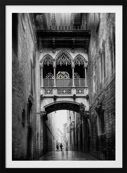 A moody, vertical black and white photograph of an ornate Gothic-style bridge connecting two stone buildings over a narrow alleyway. Below the arch of the bridge, the wet street stretches into the distance where two figures are walking away. Decor