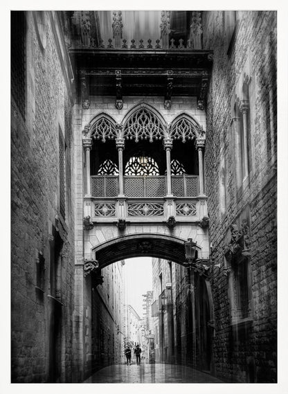 A moody, vertical black and white photograph of an ornate Gothic-style bridge connecting two stone buildings over a narrow alleyway. Below the arch of the bridge, the wet street stretches into the distance where two figures are walking away. Decor