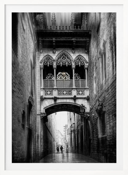 A moody, vertical black and white photograph of an ornate Gothic-style bridge connecting two stone buildings over a narrow alleyway. Below the arch of the bridge, the wet street stretches into the distance where two figures are walking away. Decor