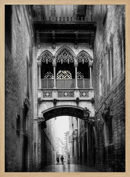 A moody, vertical black and white photograph of an ornate Gothic-style bridge connecting two stone buildings over a narrow alleyway. Below the arch of the bridge, the wet street stretches into the distance where two figures are walking away. Decor