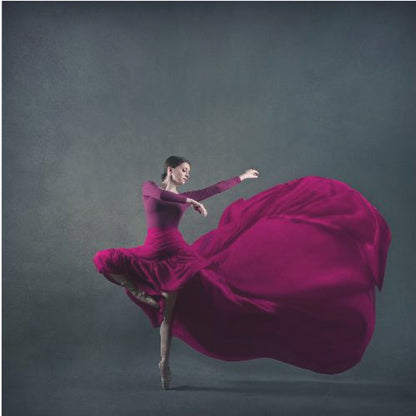 A graceful ballet dancer in a vibrant magenta dress strikes a pose on pointe, with the long, flowing fabric of her skirt billowing out dramatically behind her against a dark, textured gray background, all enclosed in a simple light wood frame. Decor