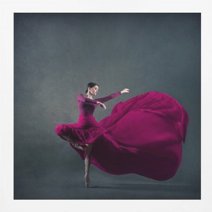 A graceful ballet dancer in a vibrant magenta dress strikes a pose on pointe, with the long, flowing fabric of her skirt billowing out dramatically behind her against a dark, textured gray background, all enclosed in a simple light wood frame. Decor