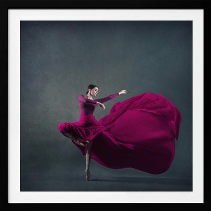 A graceful ballet dancer in a vibrant magenta dress strikes a pose on pointe, with the long, flowing fabric of her skirt billowing out dramatically behind her against a dark, textured gray background, all enclosed in a simple light wood frame. Decor