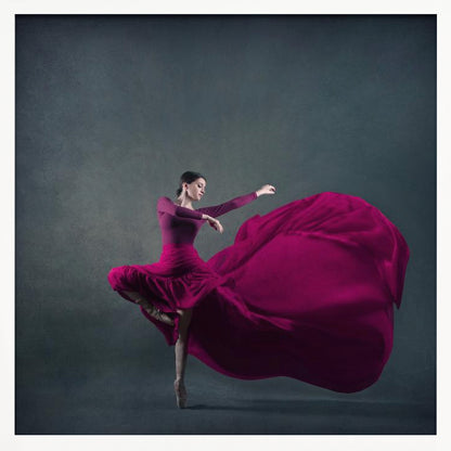 A graceful ballet dancer in a vibrant magenta dress strikes a pose on pointe, with the long, flowing fabric of her skirt billowing out dramatically behind her against a dark, textured gray background, all enclosed in a simple light wood frame. Decor