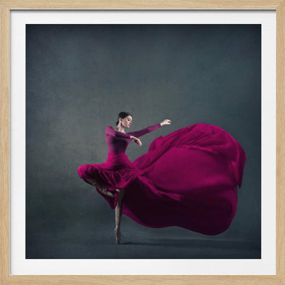 A graceful ballet dancer in a vibrant magenta dress strikes a pose on pointe, with the long, flowing fabric of her skirt billowing out dramatically behind her against a dark, textured gray background, all enclosed in a simple light wood frame. Decor