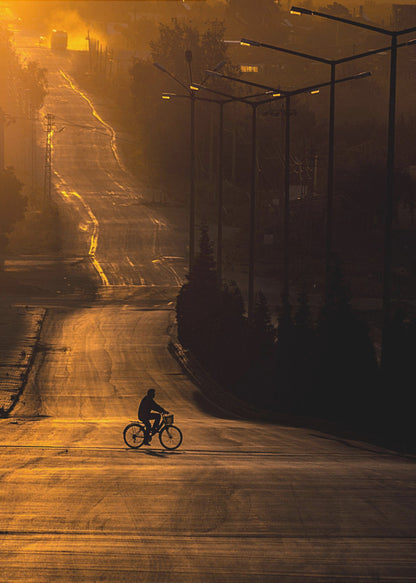 A silhouette of a person riding a bicycle across a wide, winding road illuminated by the warm, golden light of a sunrise or sunset. The road stretches uphill into the distance, and tall streetlights stand in shadow on the right. Artwork