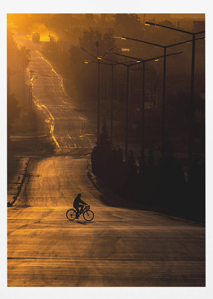 A silhouette of a person riding a bicycle across a wide, winding road illuminated by the warm, golden light of a sunrise or sunset. The road stretches uphill into the distance, and tall streetlights stand in shadow on the right. Artwork