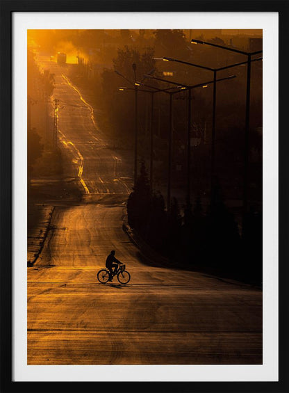 A silhouette of a person riding a bicycle across a wide, winding road illuminated by the warm, golden light of a sunrise or sunset. The road stretches uphill into the distance, and tall streetlights stand in shadow on the right. Artwork