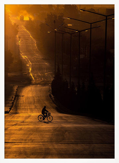 A silhouette of a person riding a bicycle across a wide, winding road illuminated by the warm, golden light of a sunrise or sunset. The road stretches uphill into the distance, and tall streetlights stand in shadow on the right. Artwork