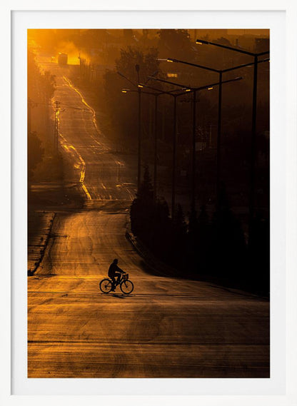 A silhouette of a person riding a bicycle across a wide, winding road illuminated by the warm, golden light of a sunrise or sunset. The road stretches uphill into the distance, and tall streetlights stand in shadow on the right. Artwork