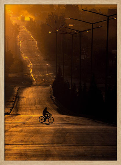 A silhouette of a person riding a bicycle across a wide, winding road illuminated by the warm, golden light of a sunrise or sunset. The road stretches uphill into the distance, and tall streetlights stand in shadow on the right. Artwork