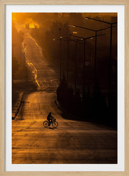 A silhouette of a person riding a bicycle across a wide, winding road illuminated by the warm, golden light of a sunrise or sunset. The road stretches uphill into the distance, and tall streetlights stand in shadow on the right. Artwork