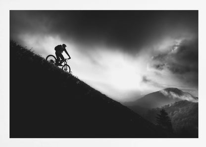 A striking black and white photo of a mountain biker's silhouette on a steep decline, set against a dramatic, light-filled cloudy sky with distant, misty mountains. Artwork