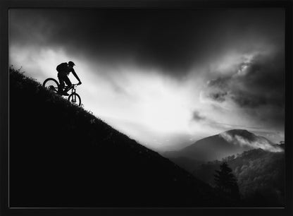 A striking black and white photo of a mountain biker's silhouette on a steep decline, set against a dramatic, light-filled cloudy sky with distant, misty mountains. Artwork