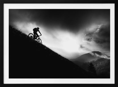 A striking black and white photo of a mountain biker's silhouette on a steep decline, set against a dramatic, light-filled cloudy sky with distant, misty mountains. Artwork