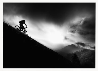 A striking black and white photo of a mountain biker's silhouette on a steep decline, set against a dramatic, light-filled cloudy sky with distant, misty mountains. Artwork