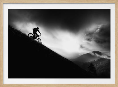A striking black and white photo of a mountain biker's silhouette on a steep decline, set against a dramatic, light-filled cloudy sky with distant, misty mountains. Artwork