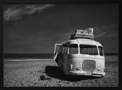 A dramatic black and white photograph of a vintage bus with luggage stacked on the roof, parked on a sandy beach. The ocean stretches out to the horizon under a dark, moody sky, with a small flock of birds flying in the distance. Print