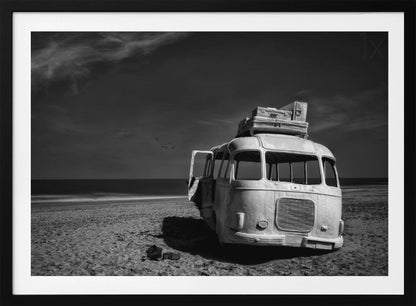 A dramatic black and white photograph of a vintage bus with luggage stacked on the roof, parked on a sandy beach. The ocean stretches out to the horizon under a dark, moody sky, with a small flock of birds flying in the distance. Print