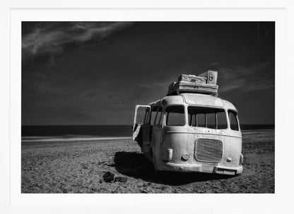 A dramatic black and white photograph of a vintage bus with luggage stacked on the roof, parked on a sandy beach. The ocean stretches out to the horizon under a dark, moody sky, with a small flock of birds flying in the distance. Print