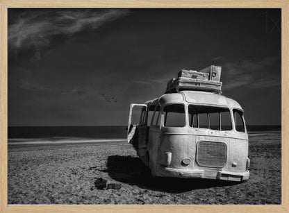 A dramatic black and white photograph of a vintage bus with luggage stacked on the roof, parked on a sandy beach. The ocean stretches out to the horizon under a dark, moody sky, with a small flock of birds flying in the distance. Print