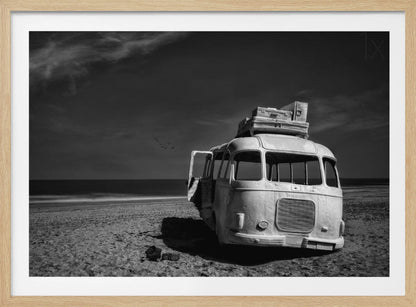 A dramatic black and white photograph of a vintage bus with luggage stacked on the roof, parked on a sandy beach. The ocean stretches out to the horizon under a dark, moody sky, with a small flock of birds flying in the distance. Print