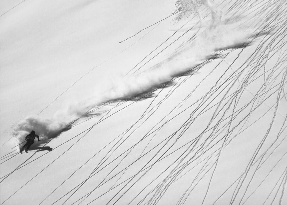 A dramatic black and white overhead photograph of a skier carving down a steep, powder-filled mountain slope, kicking up a huge plume of snow. Numerous ski tracks crisscross the pristine white snow, creating a pattern of dark lines. Poster